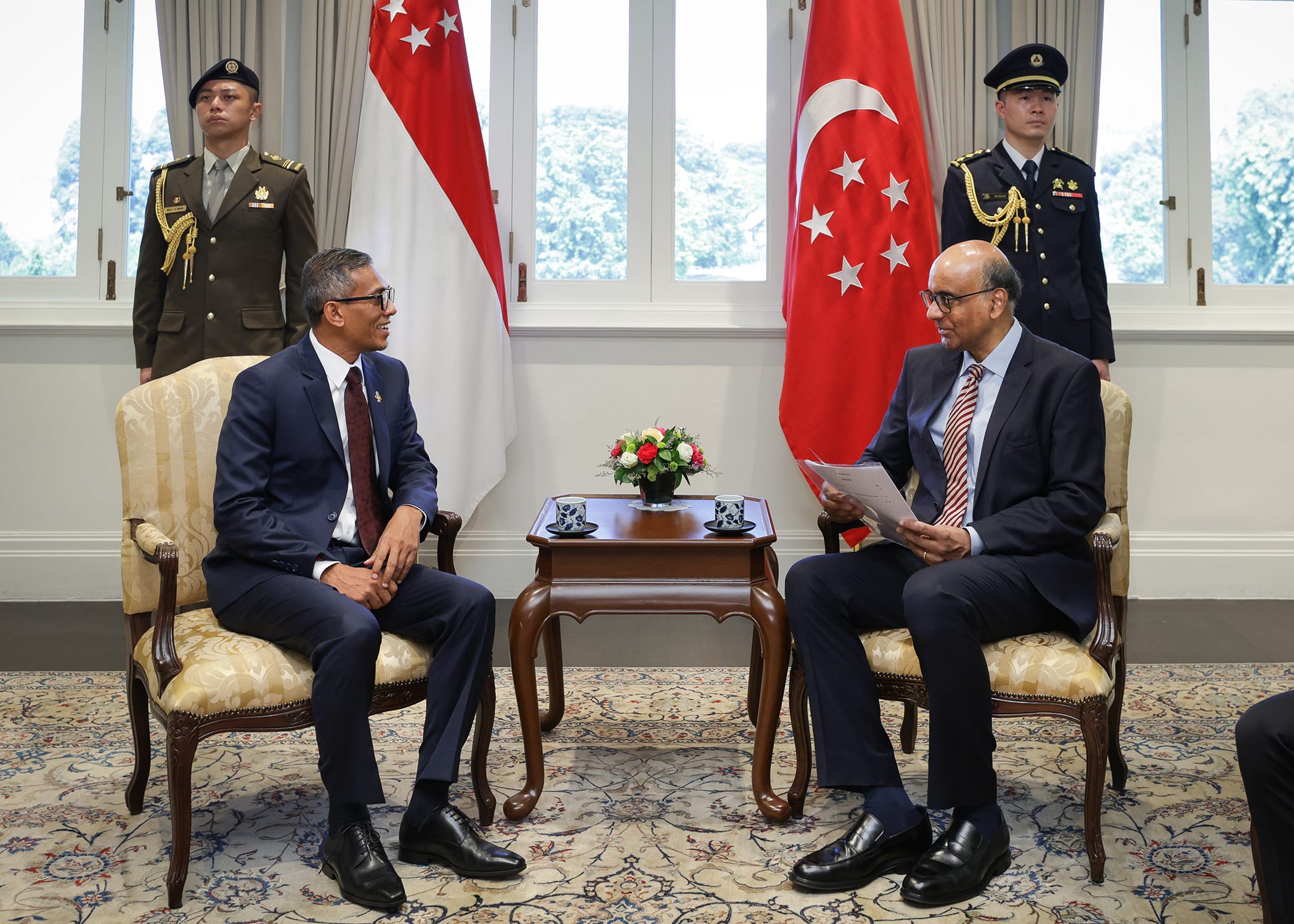 Two men in suits seated, facing each other, with Singapore flags and uniformed guards in the background.